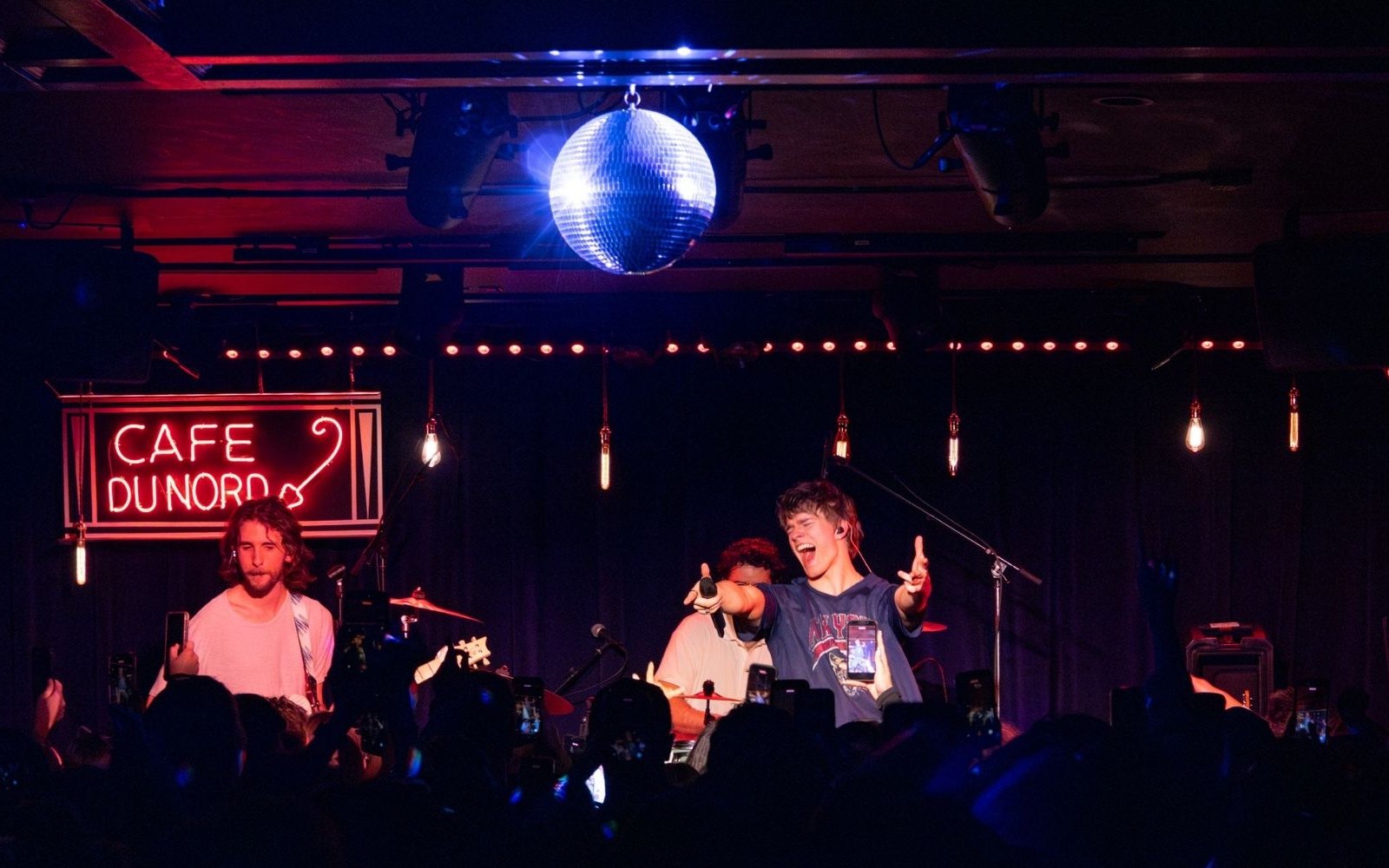 A singer pointing out at the crowd on the Cafe du Nord stage, under the neon sign and a disco ball, with phones held up in front